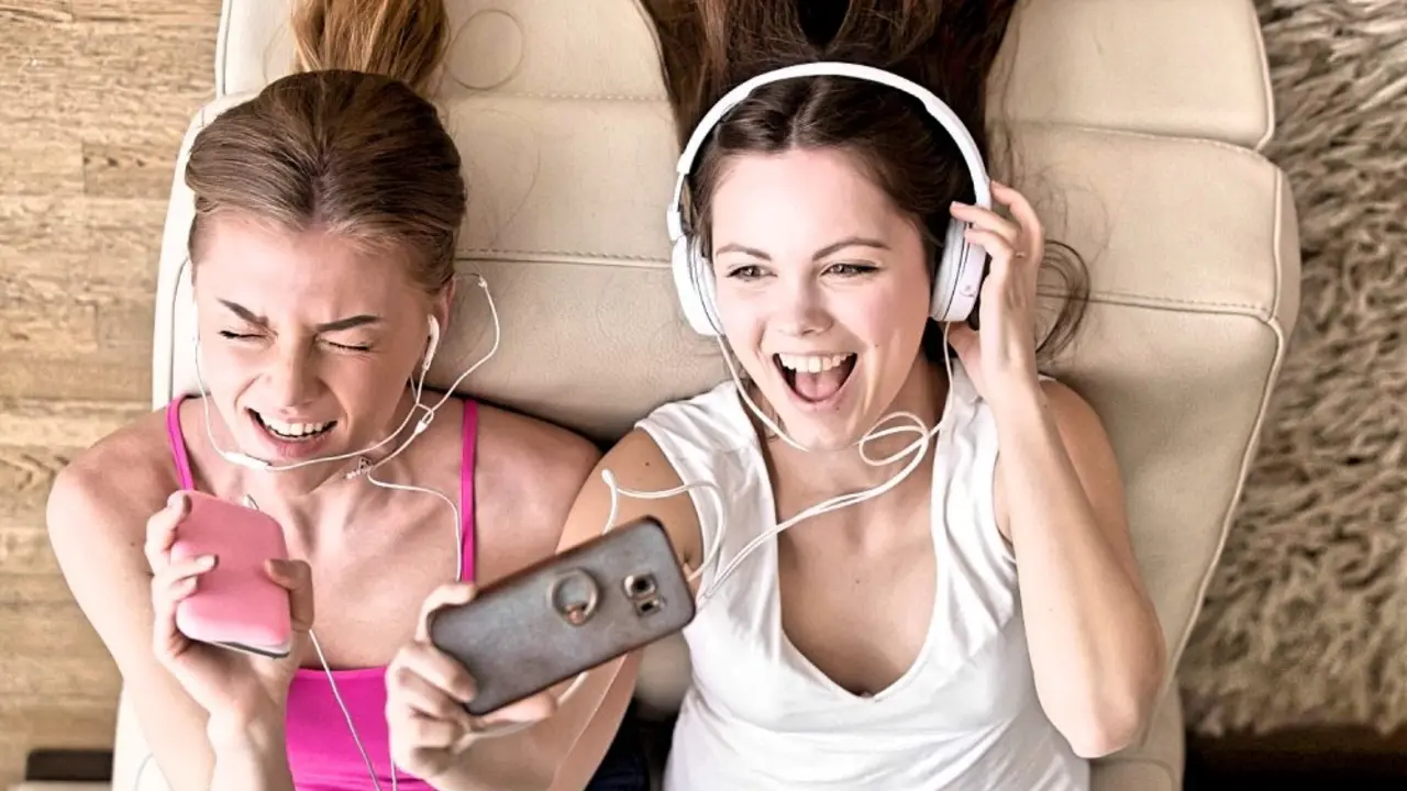 Close up portrait of two excited girlfriends enjoying music in headphones, making selfie photo with cellphone while lying on sofa at home. Teen girls having fun, singing popular hit together. Top view