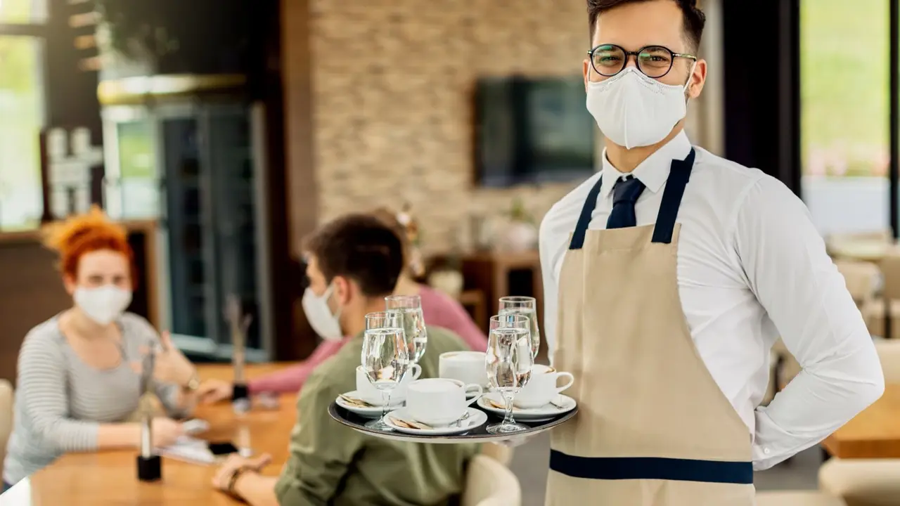Happy waiter wearing protective face mask while serving coffee to guests during coronavirus epidemic. 